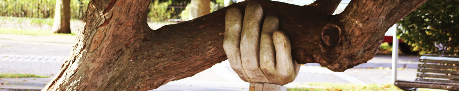 Ein dicker Baumstamm, der schräg gewachsen ist, wird von einer aus Holz geschnitzen Hand gestützt.